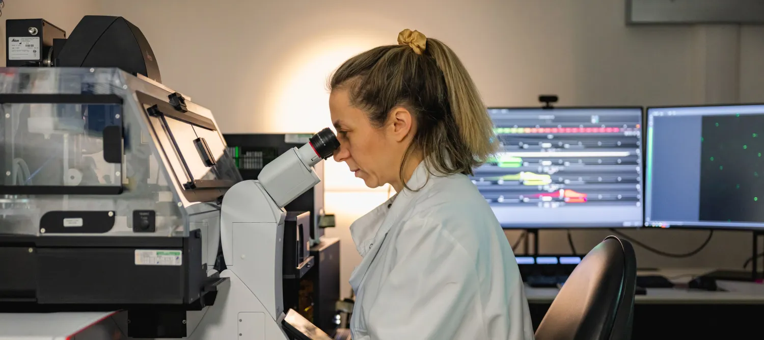 Researcher looks down microscope in a dimmed room with a lit computer screen in the background