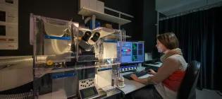Scientist at computer next to microsope in dark room