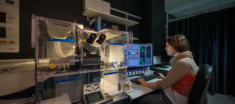 Scientist at computer next to microsope in dark room