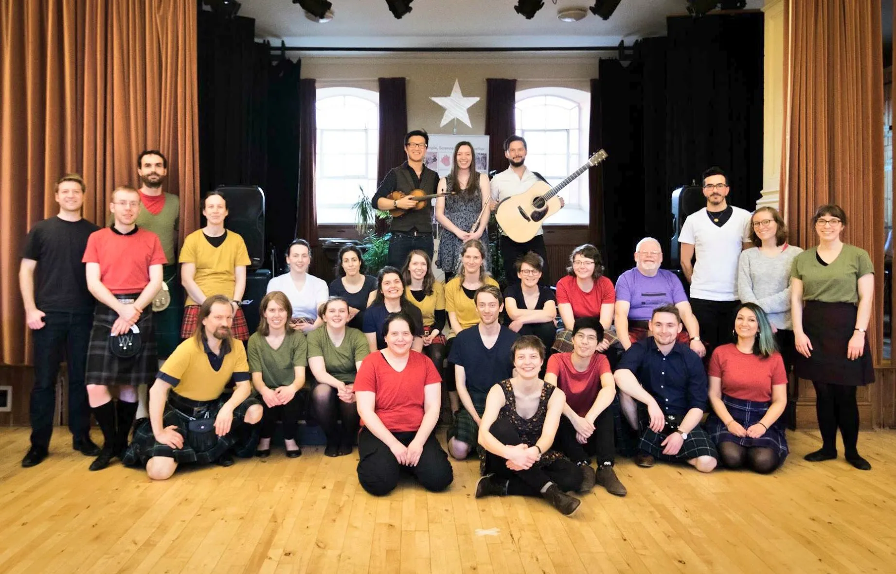 A group of ceilidh dancers in a local community hall, posing with the ceilidh band in the background.