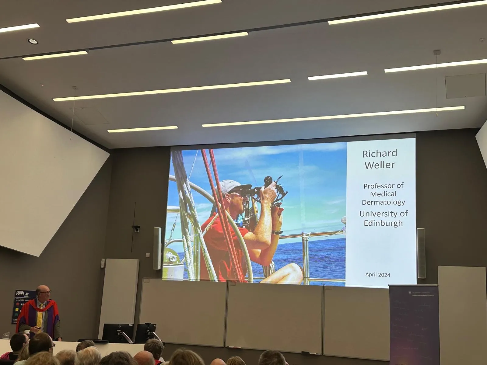 An audience looking at a projector screen showing an old photograph of a professor on a boat using a sextant