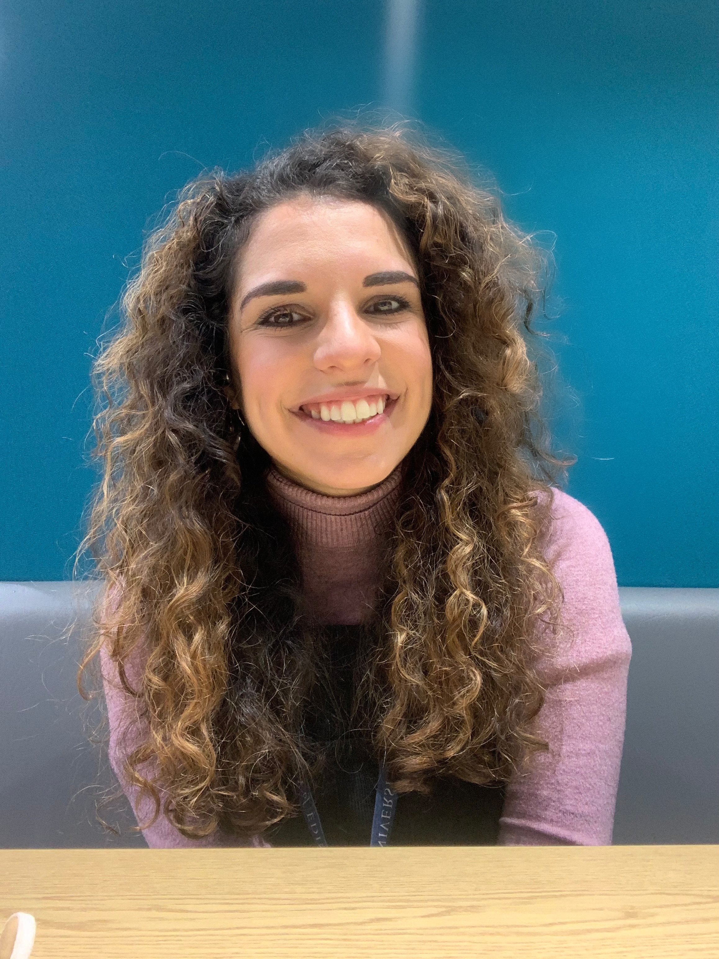 A woman with curly brown hair and a pink jumper smiles at the camera