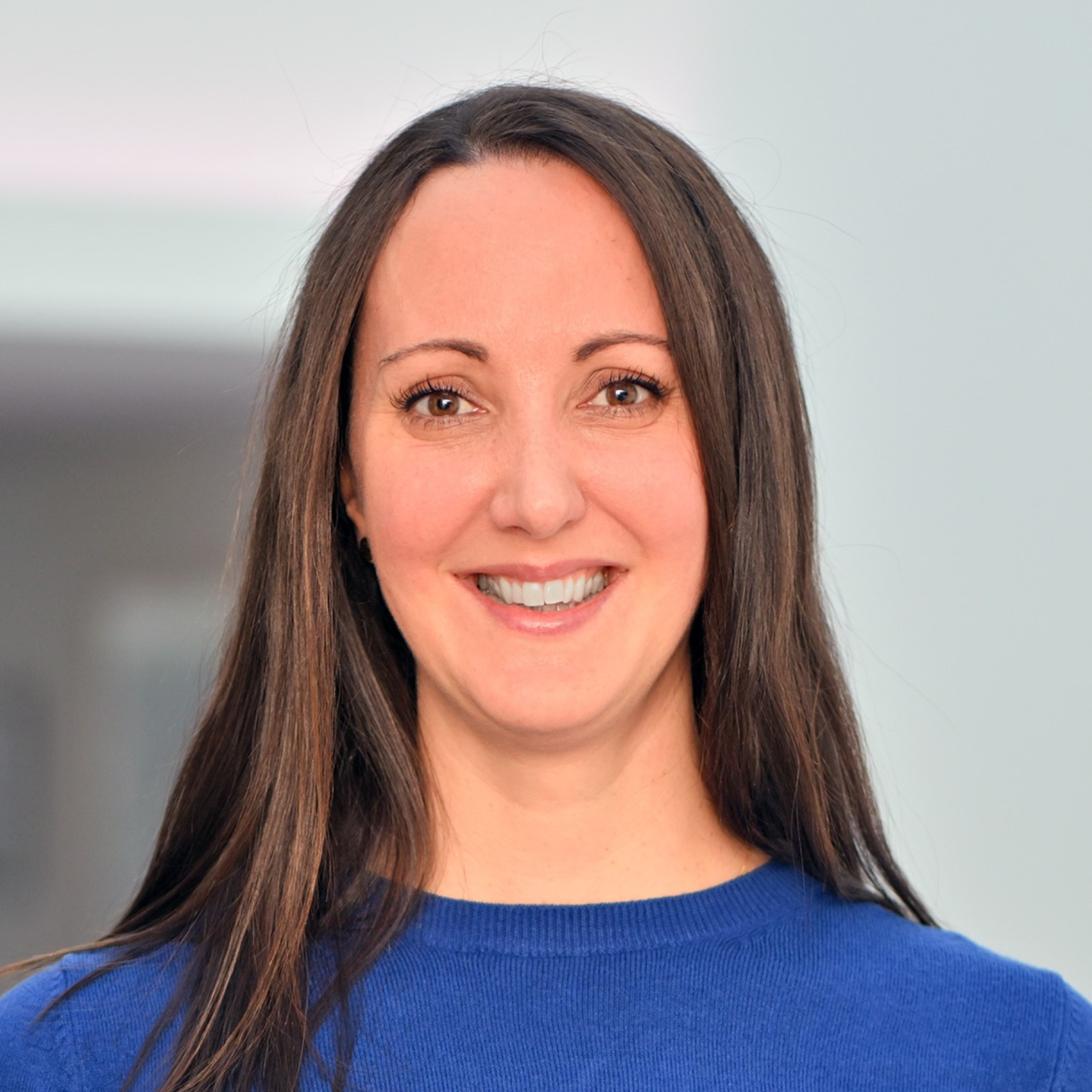 a portrait photograph of Chloe Stanton, a white woman with long dark brown hair, wearing a royal blue jumper