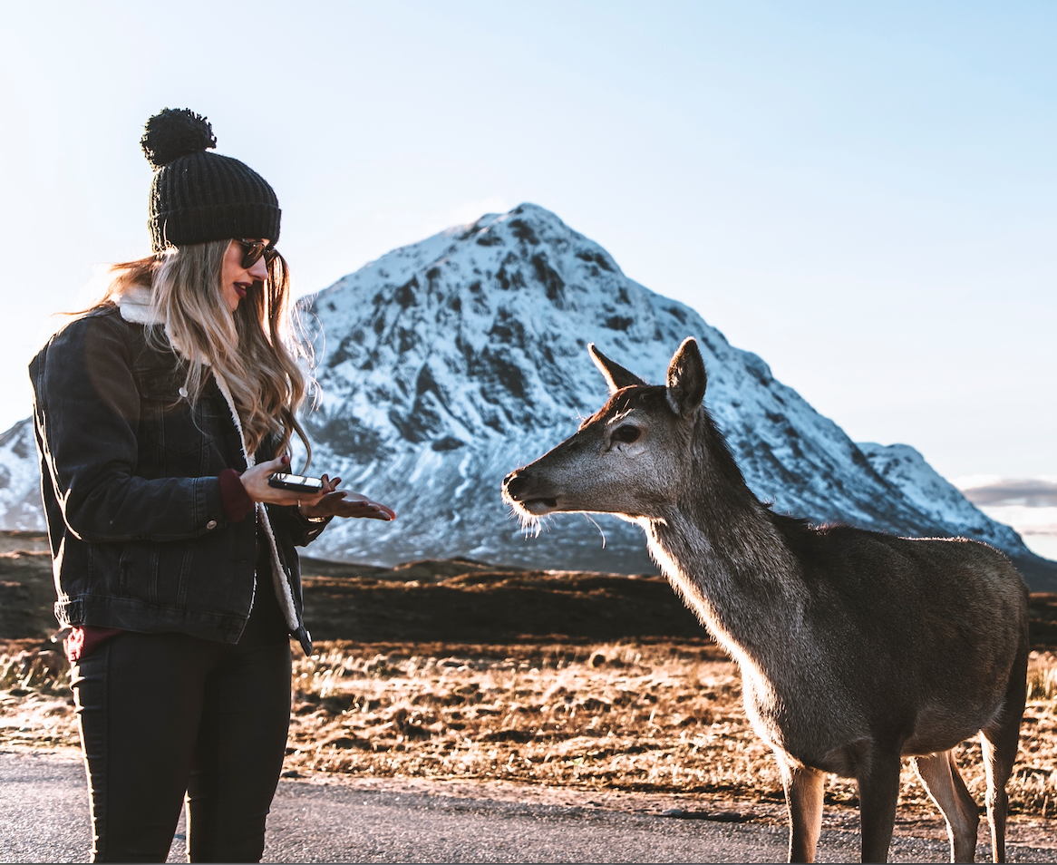 A woman next to a female deer, in front of a snow covered mountain