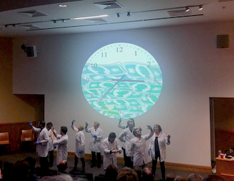 A group of performers inspecting Petri dishes at the front of an auditorium